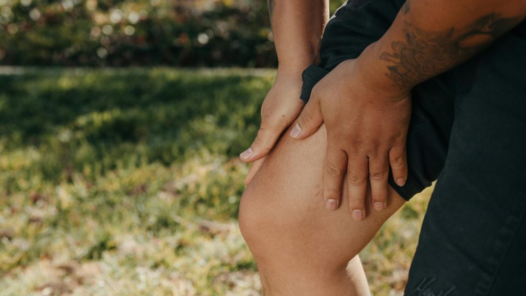 Close-up of a man's hands gripping his painful knee outdoors, with a grassy background.