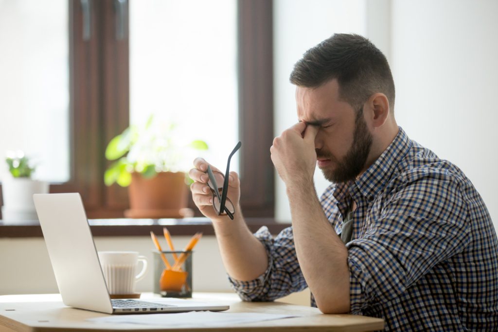 Man sitting at a laptop holding his glasses and pressing his fingers to the bridge of his nose due to a headache from prolonged screen use