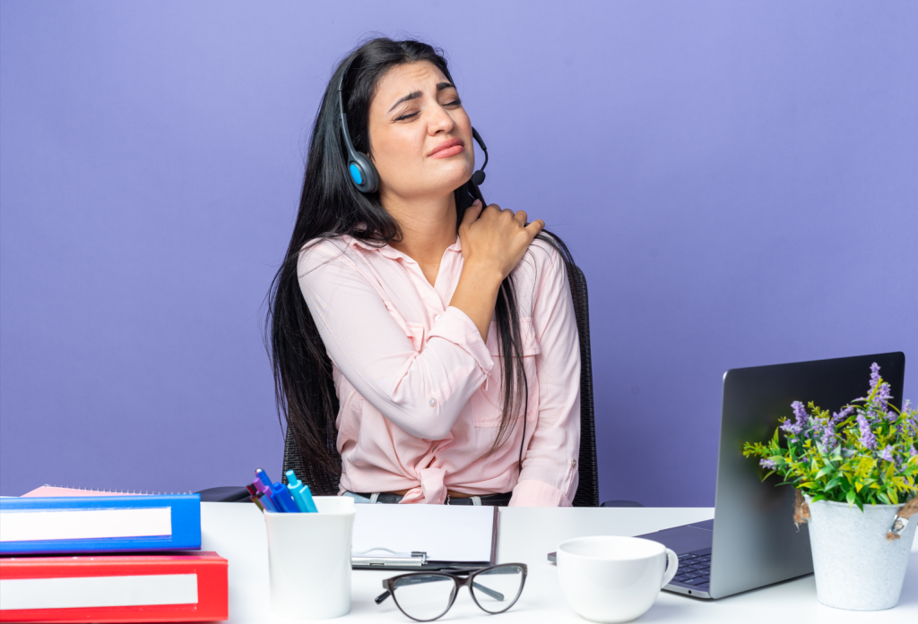 Young woman holding her shoulder in pain while working on a laptop, a common sign of early cervical spondylosis