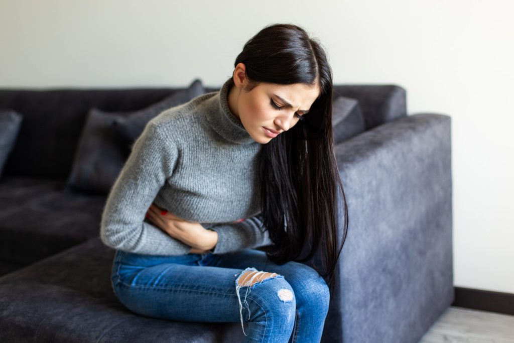 Woman sitting on a sofa holding her stomach in pain, a common symptom linked to navel displacement