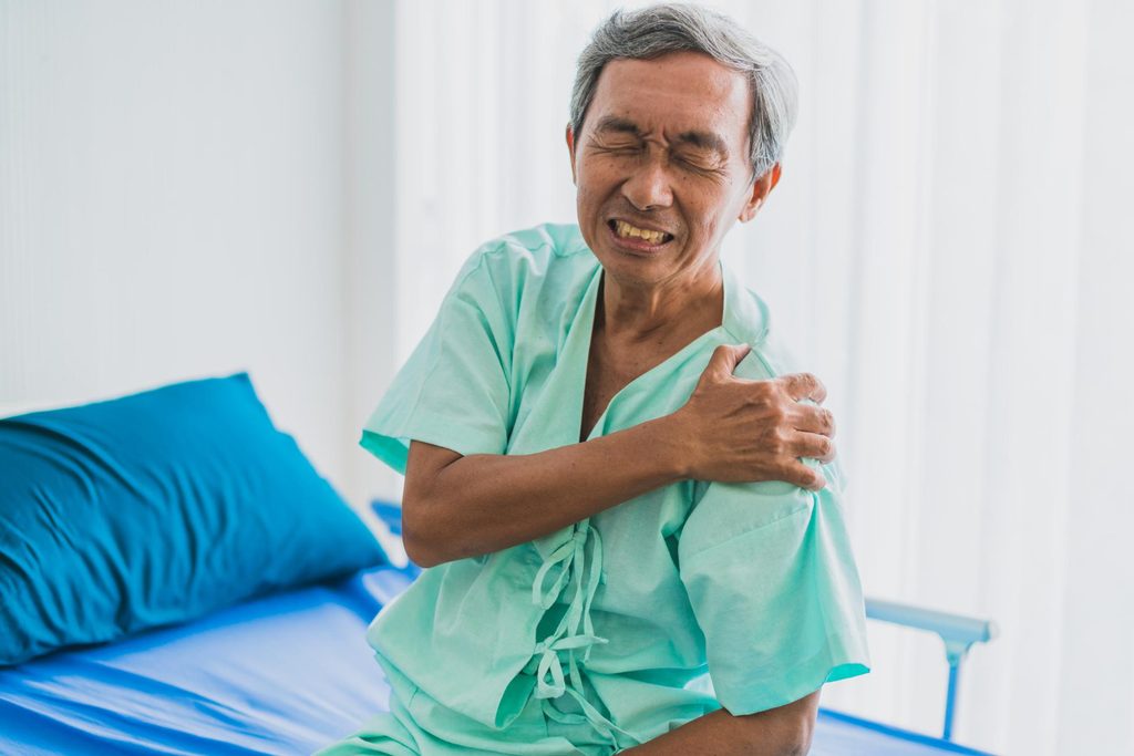 Older man in a hospital gown wincing and gripping his left shoulder, showing the pain of frozen shoulder